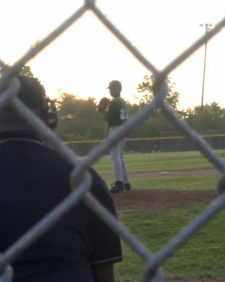 A dark, backlit phone photo of a pitcher, taken through the backstop fence