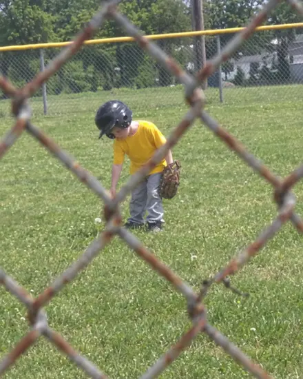 A far-away phone photo of a small boy in the outfield, seen through a rusty fence